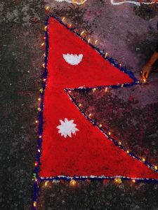 A close-up of a Nepali flag-shaped decoration with white floral designs, surrounded by glowing candles, as a hand places one on the textured, colorful surface.