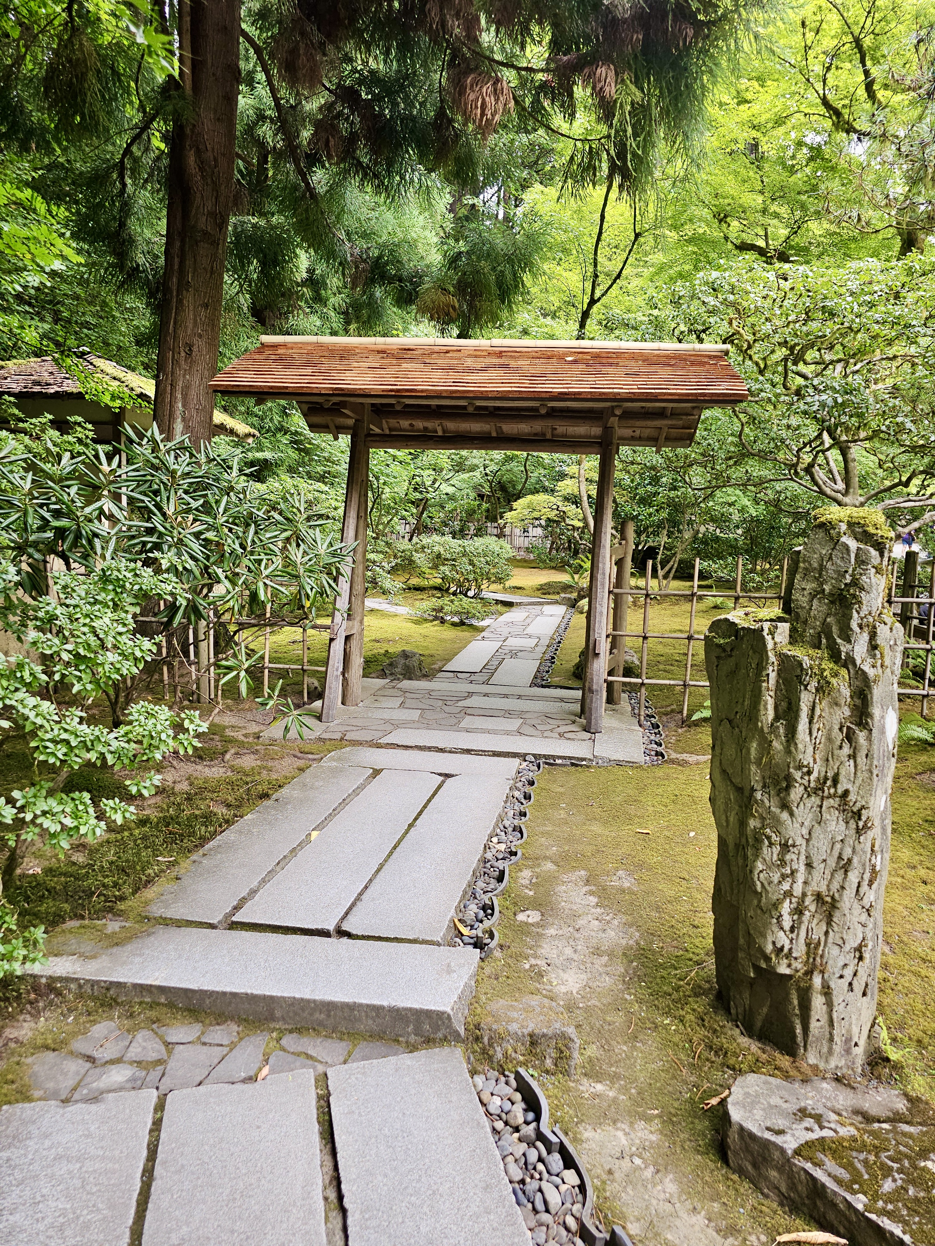 A wooden garden gate with a stone path leading through green trees and mossy ground. Photo taken at Portland Japanese Garden, Oregon, USA. 