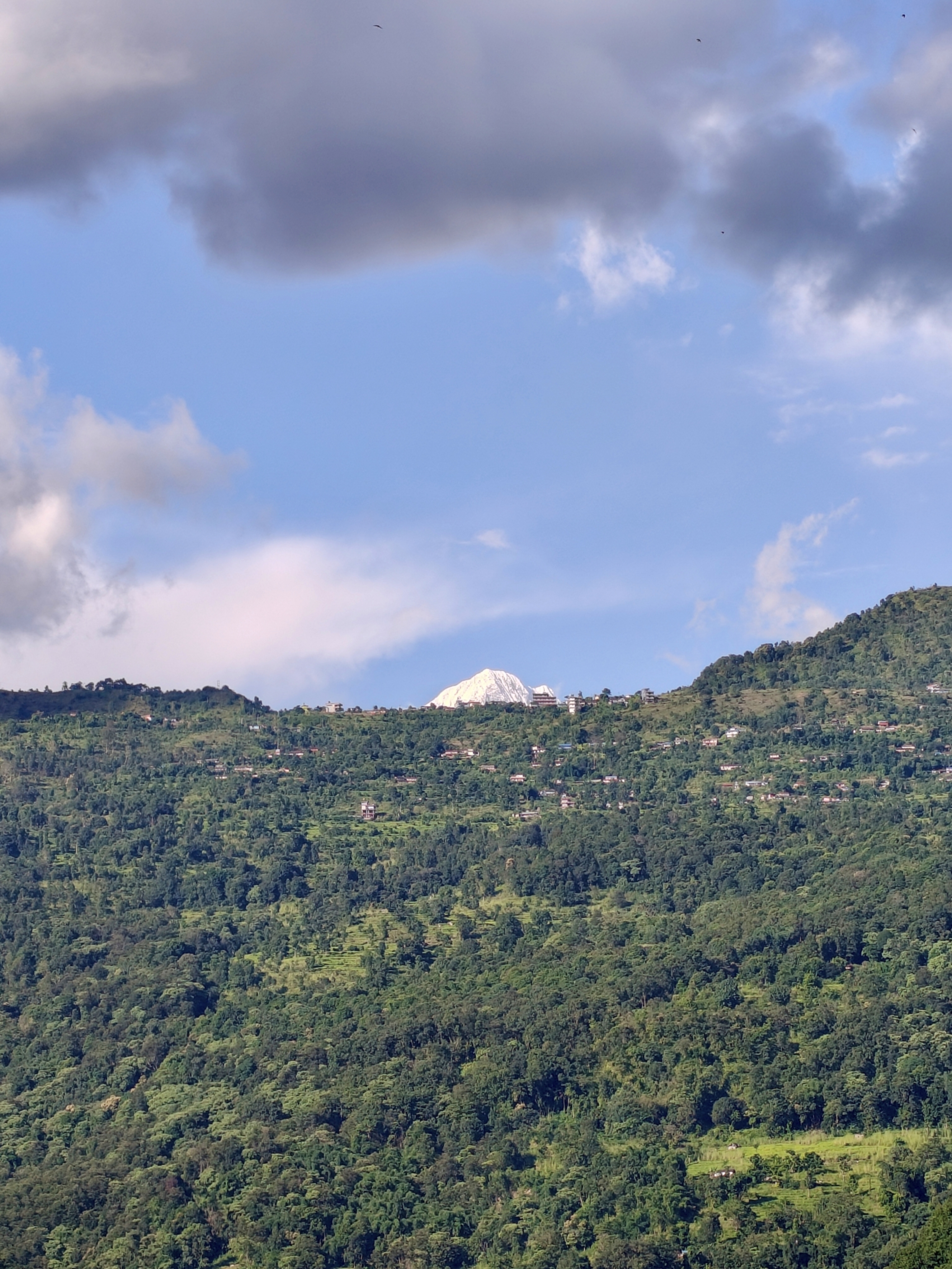 A scenic landscape featuring a lush green mountain slope dotted with small buildings, rising beneath a clear blue sky with scattered clouds
