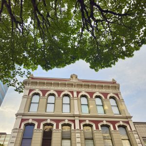A charming old building with red and cream accents in Portland Downtown, partially covered by thick green tree branches overhead.