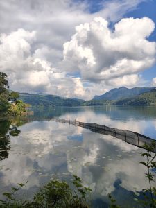 A tranquil lake scene featuring calm waters reflecting fluffy white clouds and green hills in the background. 