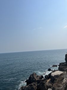 A clear blue sky stretches overhead, meeting the calm, deep blue sea at the horizon. The rocky coastline is visible in the foreground, with jagged rocks jutting out into the water. A small platform or promenade appears on the shore, where a couple of people are gathered