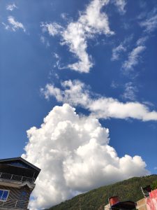 A vivid blue sky with a large white cloud over green mountains, with a patterned building and sloped roof in the foreground.