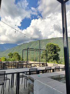 
An outdoor dining area with black tables and chairs on stone flooring, set against green mountains under a partly cloudy sky, with string lights and a nearby wooden swing.