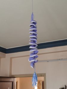 A spiral-shaped crochet decoration in shades of purple, with white edges, hanging from the ceiling