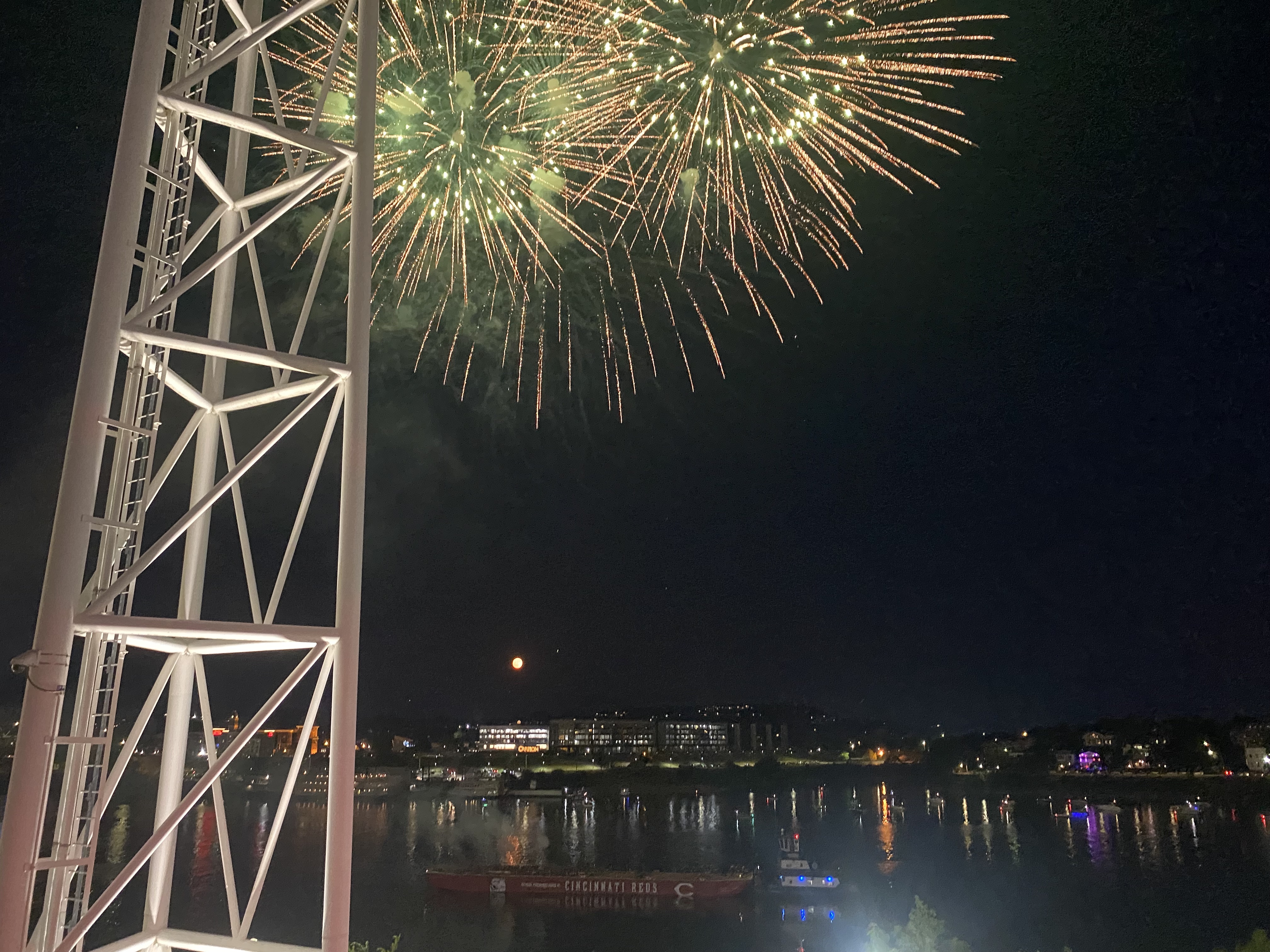 Fireworks along the Ohio River, a reflection of lights in the water. A white tower, a floating boat, and buildings are visible.