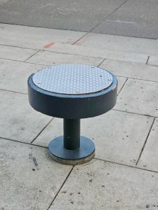A dark gray, cylindrical outdoor stool with a circular, textured metal top is seen on a paved surface. Captured from Providence Park, Portland.