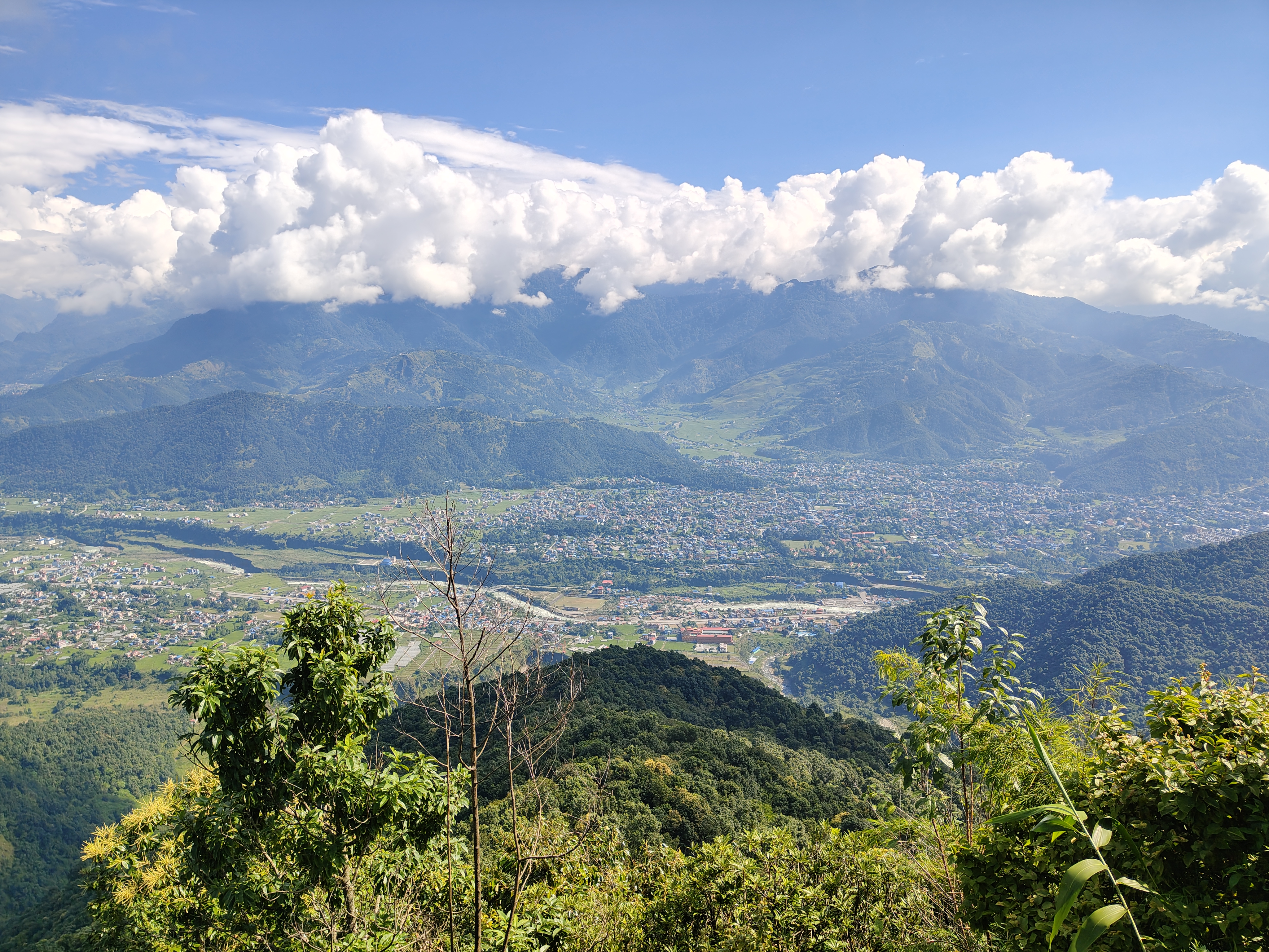 A panoramic mountain view overlooks a green valley with a winding river, towns, and fields. Rolling hills and distant mountains stretch under a blue sky with white clouds, framed by dense foreground trees.