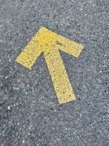 A yellow painted arrow on a rough blacktop road pointing diagonally forward, indicating direction. Photo taken near the Portland Japanese Garden, Oregon. 