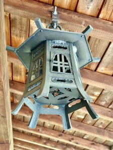 A close-up of a traditional Japanese-style hanging lantern made of metal, captured under a wooden roof at the Portland Japanese Garden. The intricate design adds cultural charm to the peaceful garden setting. 
