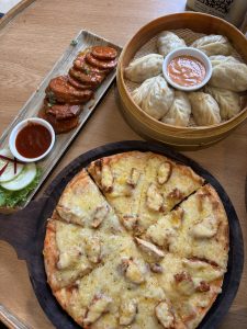 A wooden table is set with three dishes. In the foreground, a round pizza, sliced into triangular pieces, is topped with melted cheese and pieces of chicken. 