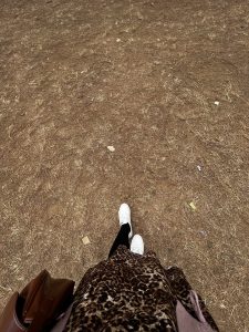 A person's legs are visible as they stand on a dry, uneven ground covered with brown dirt and scattered leaves. The individual is wearing white sneakers, black leggings, and a leopard print skirt. A brown handbag is resting against their side. The background is filled with patches of dry grass and small debris.