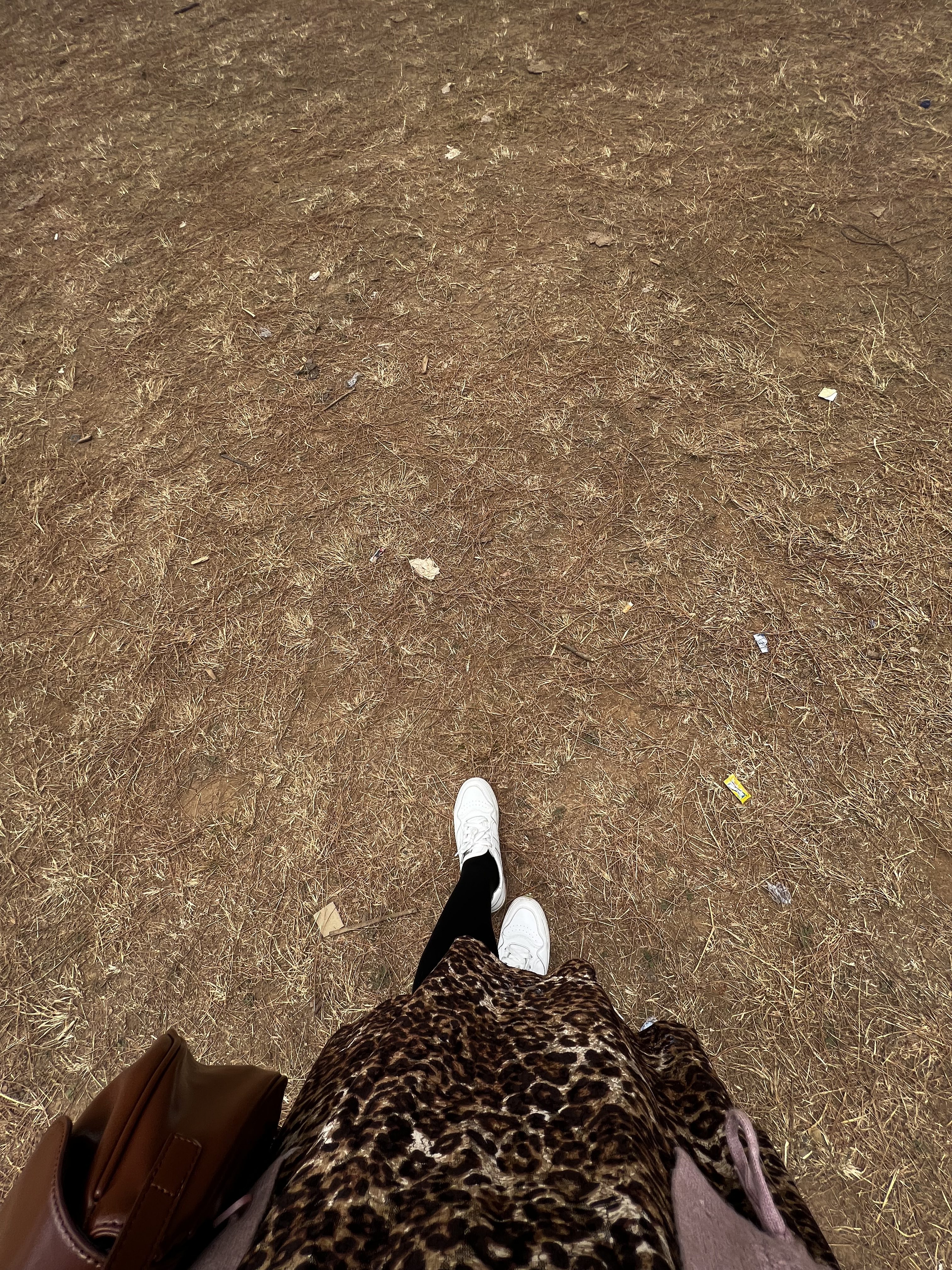A person's legs are visible as they stand on a dry, uneven ground covered with brown dirt and scattered leaves. The individual is wearing white sneakers, black leggings, and a leopard print skirt. A brown handbag is resting against their side. The background is filled with patches of dry grass and small debris.
