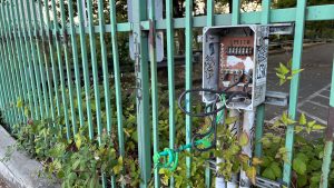 On a fence, an electrical box is shown broken open, with large electrical wires coiling out of it, cut off so the bare wire is in easy reach.