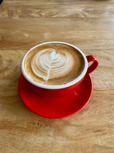 An overhead shot of a red ceramic mug and saucer on a wooden table. The mug holds a cappuccino with a beautiful, intricate heart-shaped art made in the milk foam.