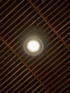 A close-up of a single decorative ceiling light at Portland International Airport. The lamp has a sunburst-style design and hangs from a wooden slat ceiling, adding an artistic touch to the airport architecture. 
