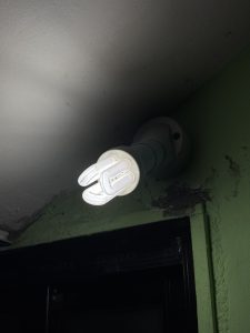A ceiling-mounted light fixture with a compact fluorescent bulb, illuminating a portion of the wall and ceiling.