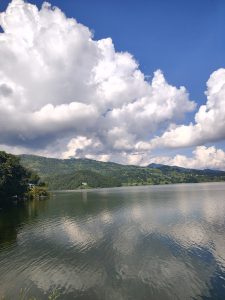 A tranquil landscape featuring a calm lake reflecting fluffy white clouds and a blue sky. 