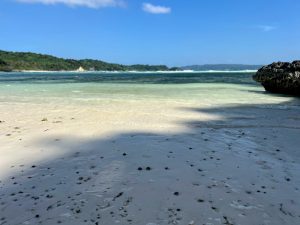 A wide shot of a white sand beach with gentle waves lapping the shore and a green, tree-covered hill in the background.