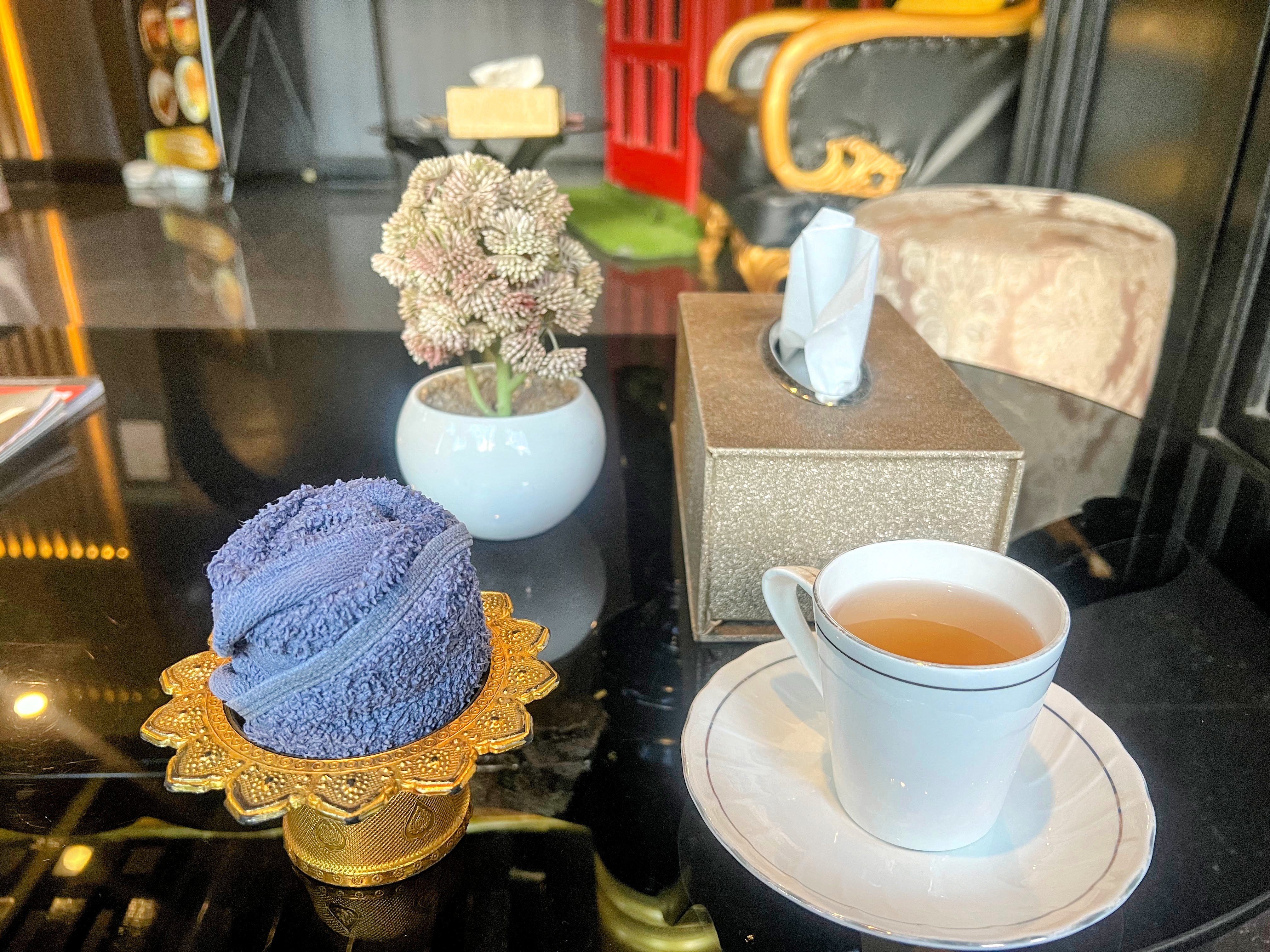 A small cup of tea, a rolled-up towel, a tissue box, and a potted plant are arranged on a shiny black table.