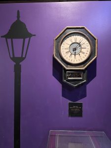 An antique octagonal wall clock hangs on a vibrant purple wall next to the black silhouette of a Victorian-style street lamp.
