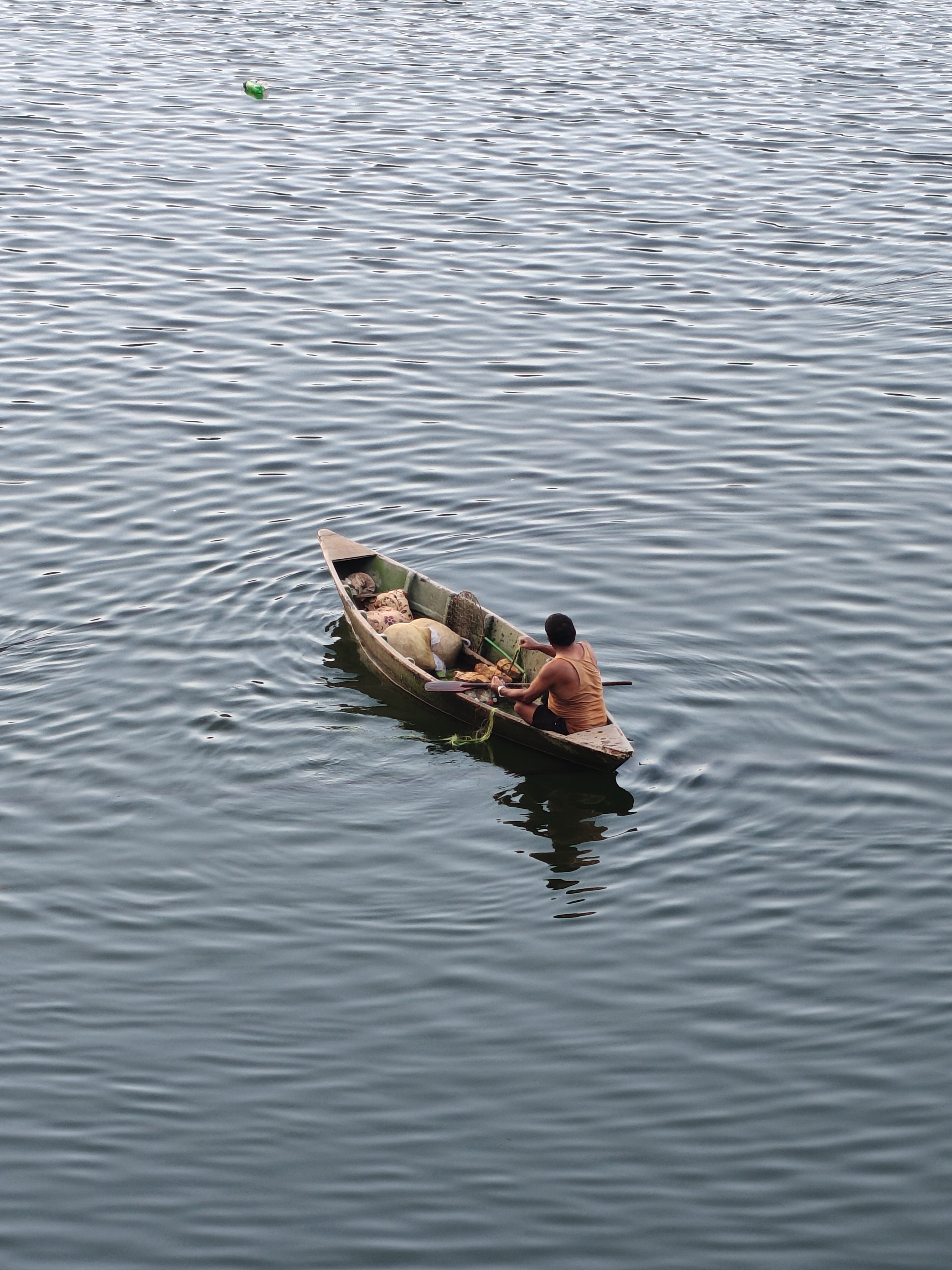 A small wooden boat carrying a lone person and fishing gear drifts on calm, rippling water, with a green object visible in the distance.