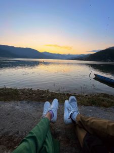 Two pairs of legs in white sneakers resting near the edge of a Fewa lake at sunset, with mountains silhouetted against the orange and blue sky in the background.