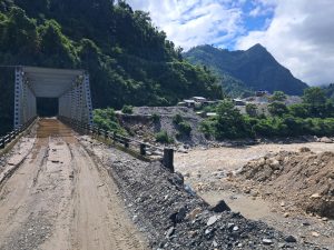 A muddy dirt road leading to a metal bridge over a rocky river, surrounded by lush green hills and a small village with houses nestled among the trees, under a partly cloudy sky.