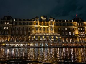 A grand riverside building lit with golden lights at night, its many windows and arched doorways reflecting warmly on the rippling water below. The elegant architecture features multiple stories, decorative details, and a central glass atrium that glows brightly.