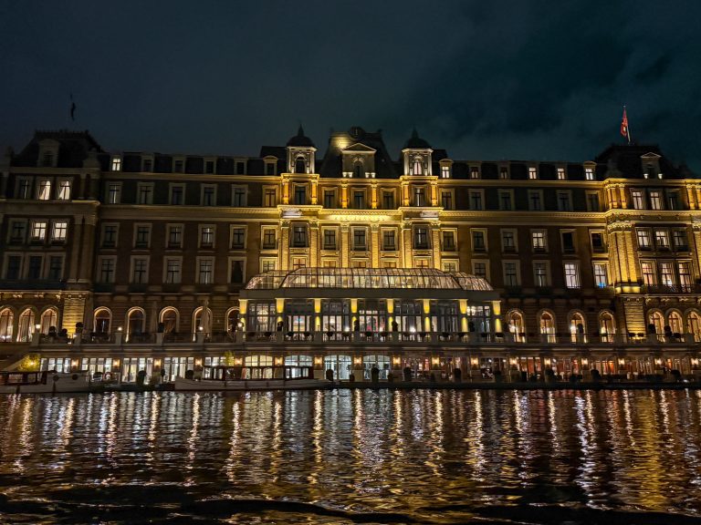 A grand riverside building lit with golden lights at night, its many windows and arched doorways reflecting warmly on the rippling water below. The elegant architecture features multiple stories, decorative details, and a central glass atrium that glows brightly.