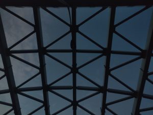 A geometric roof structure viewed from below, with intersecting metal beams forming a triangular grid against a dusk sky.