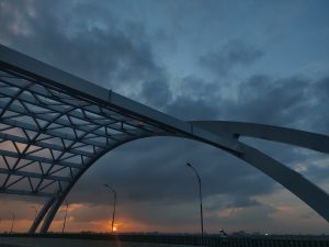 A modern arch bridge with a geometric design is silhouetted against a dramatic sky at sunset.