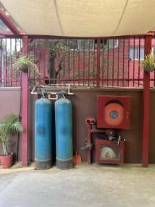 An outdoor plumbing and a fire hose reel. Two large blue tanks are next to a red box containing a fire hose. There are also two small potted plants hanging from red metal poles.