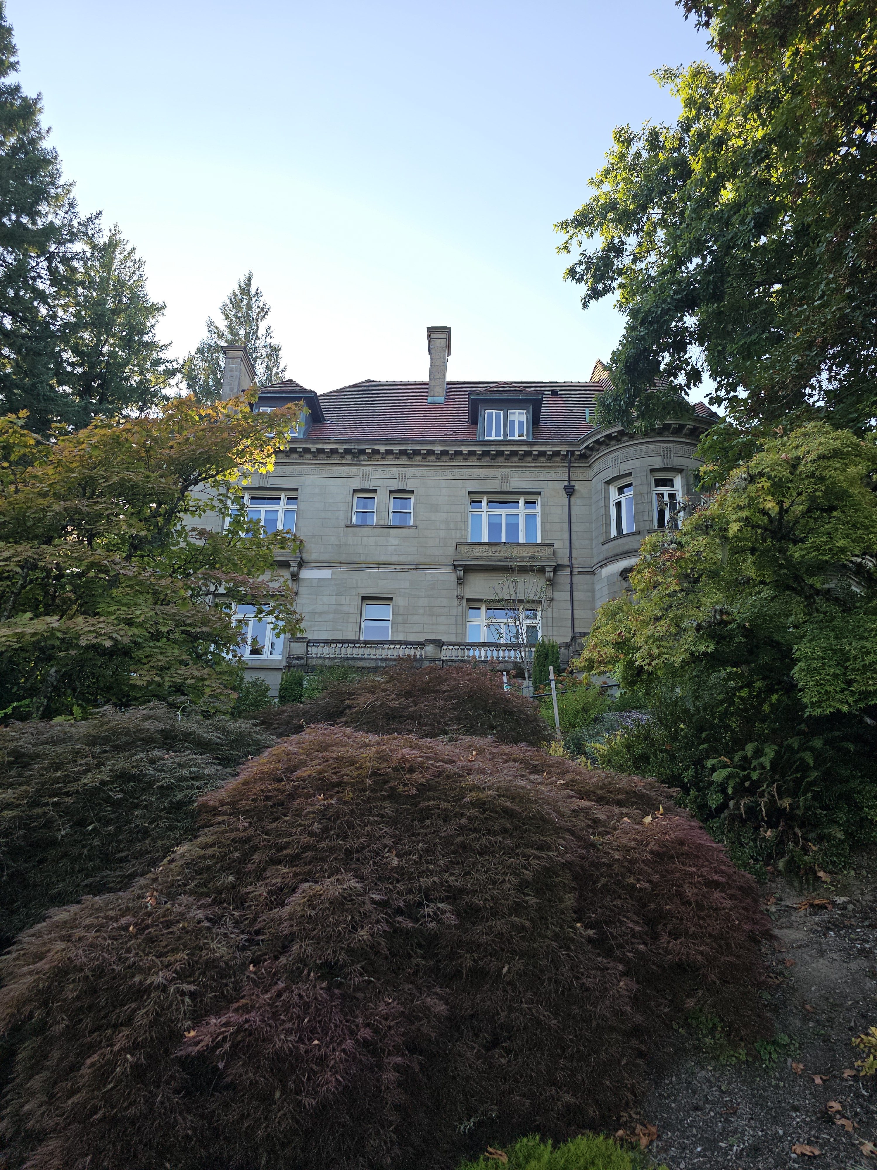 A grand historic building viewed from below, with a red roof, stone walls, and surrounded by lush trees. Photo taken at Pittock Mansion, Portland.