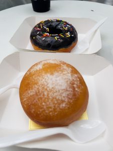Two donuts in separate white paper trays are shown, one topped with chocolate frosting and sprinkles and the other dusted with powdered sugar.