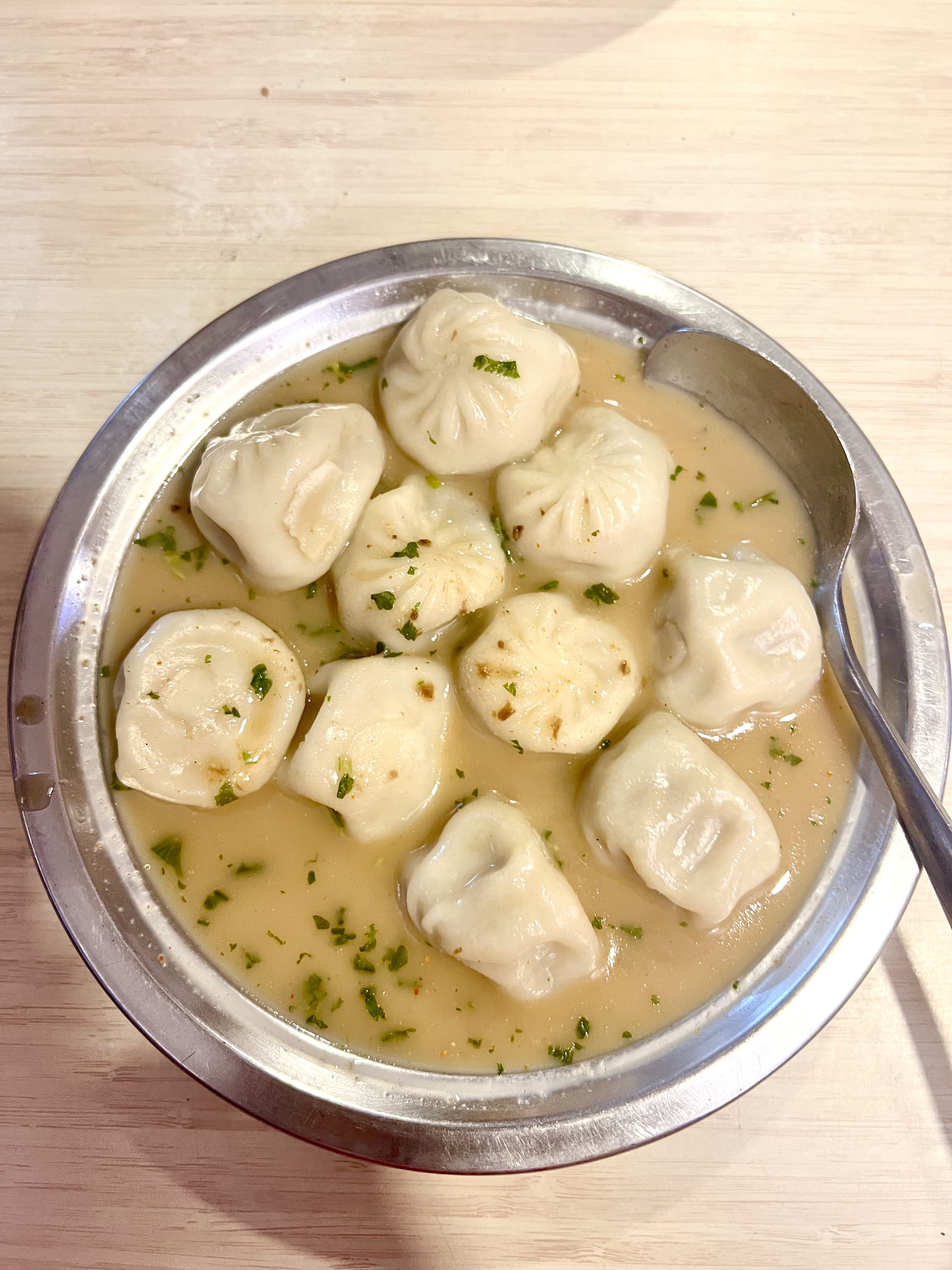 A metal plate holds several local dumplings known as Mo:Mo, in a broth, garnished with chopped herbs.