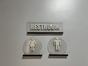 A shot of a clear, modern-style restroom sign on a light gray wall. The sign has the word "RESTROOM" on a rectangular plaque and separate circular plaques with male and female symbols underneath.