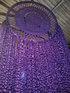 A vibrant purple dream catcher hangs from a wooden ceiling, featuring an intricate crocheted circular center surrounded by cascading fringes of purple fabric