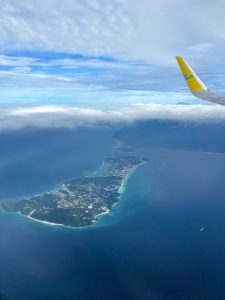 
An aerial view from an airplane window shows a long, narrow tropical island with resorts and beaches, surrounded by deep blue ocean under a blue sky partially covered by white clouds; the yellow wingtip of the plane is visible in the upper right.