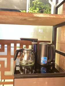 A black and silver water heater and kettle, probably used as coffee maker, are sitting on a table in front of a glass shelf.