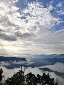
A panoramic view from above reveals green hills, a lake, and a city in the valley, with sunlight breaking through clouds and mist creating a serene, ethereal atmosphere.