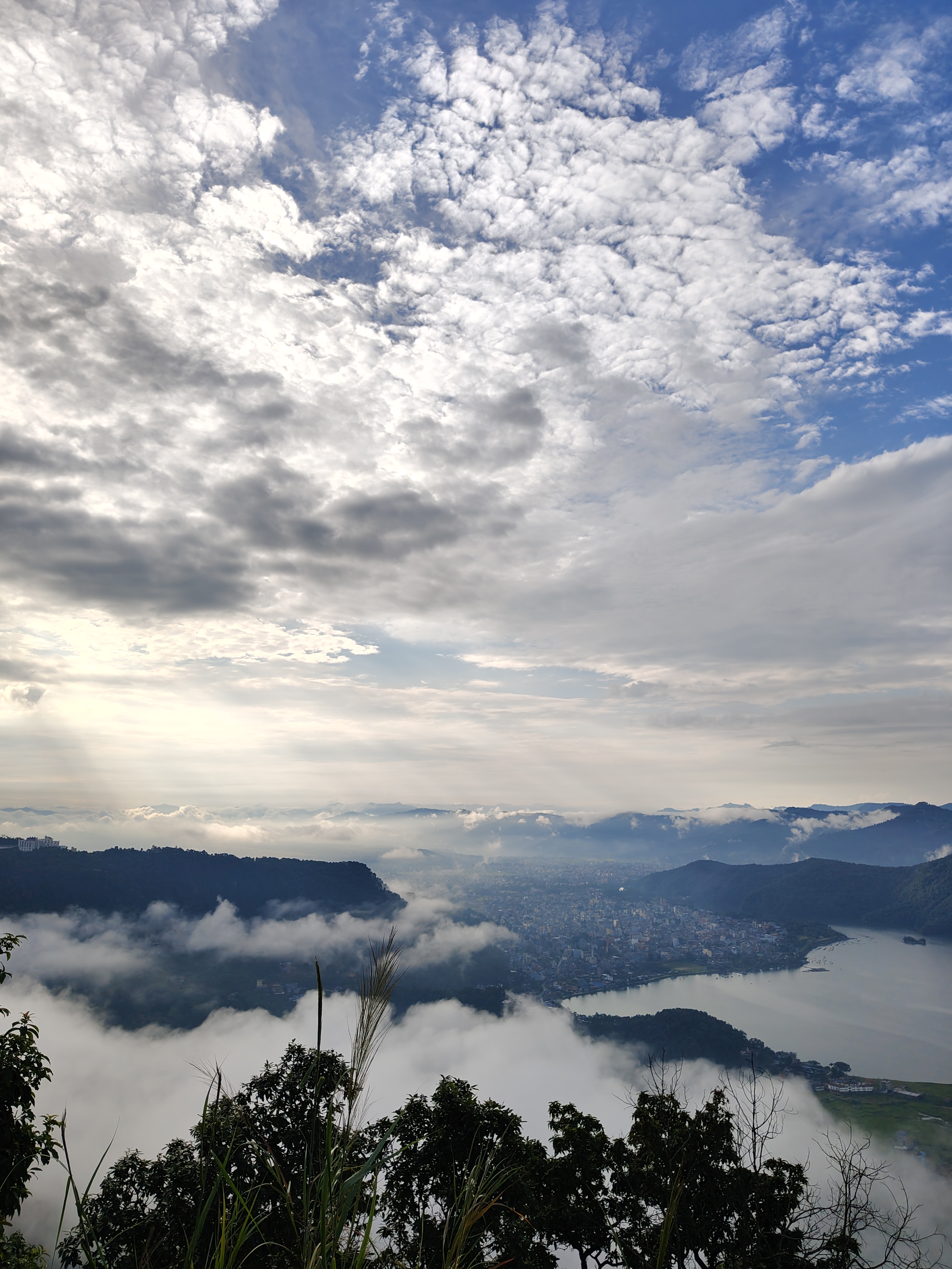 
A panoramic view from above reveals green hills, a lake, and a city in the valley, with sunlight breaking through clouds and mist creating a serene, ethereal atmosphere.