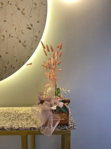 A shot of a small, decorative flower arrangement in a wooden box with a pink bow, sitting on a granite countertop. A large, circular, illuminated mirror with a leafy design reflecting on it is in the background.