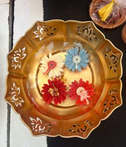 Four small, colorful flower-shaped candles floating in a water-filled, scalloped brass offering bowl.