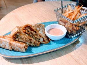 A blue plate holds chicken lachha melt with a dipping sauce, served alongside a small metal basket of seasoned French fries.