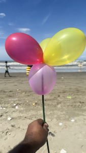 A person's hand holds up a stick with three colorful balloons at the beach, with the sand and ocean in the background.