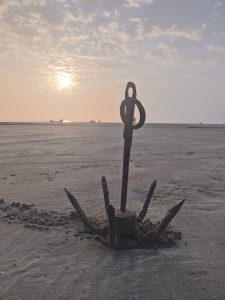 An old, rusted anchor stands upright in the sandy beach, silhouetted against a hazy sunset.