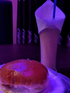 A close-up image of a sesame seed bun with a bite taken out of it, placed on a white paper. 
