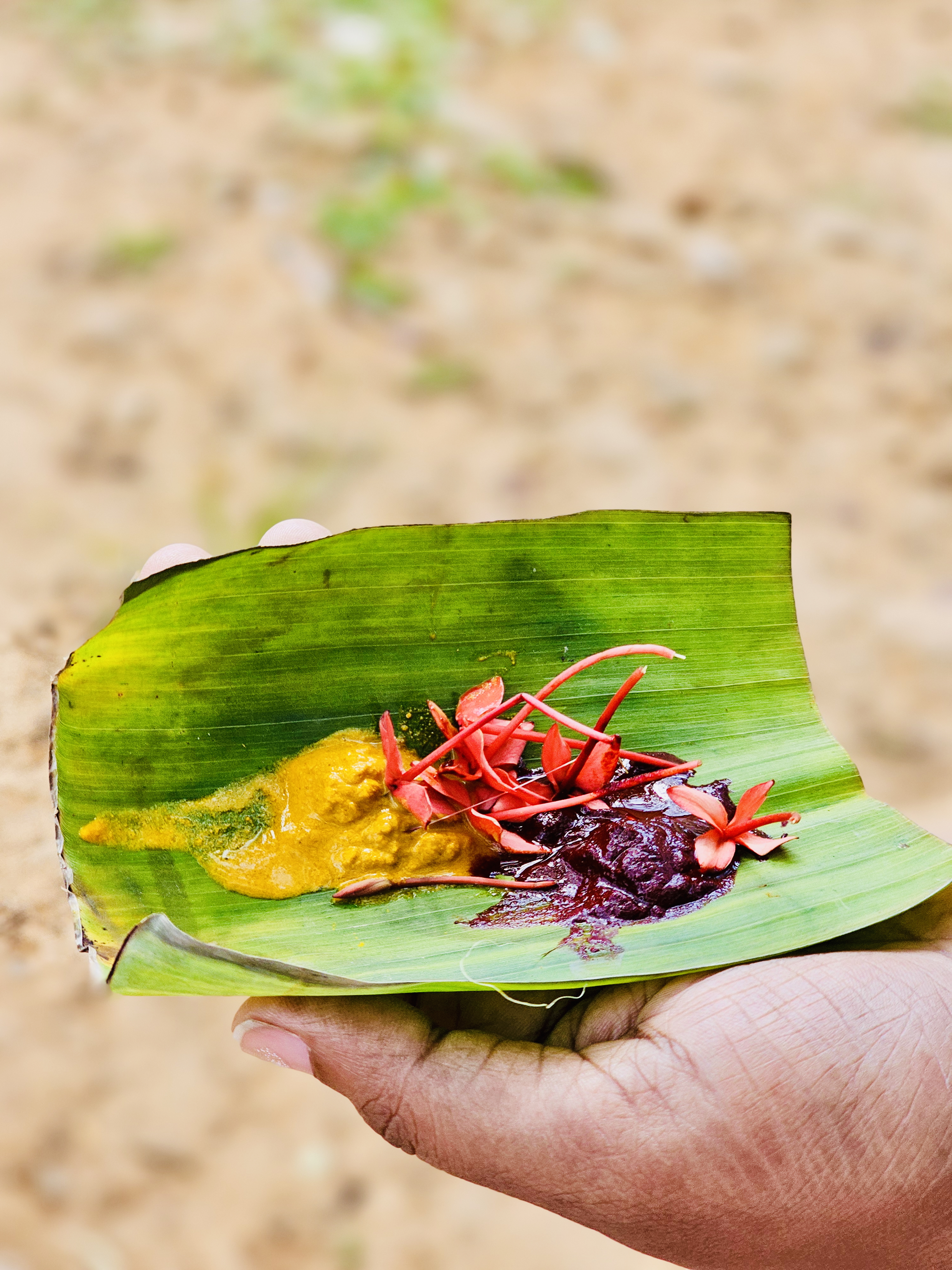 A close-up of a hand holding a banana leaf with turmeric, sandalwood paste, and red flowers, used in Kerala rituals.