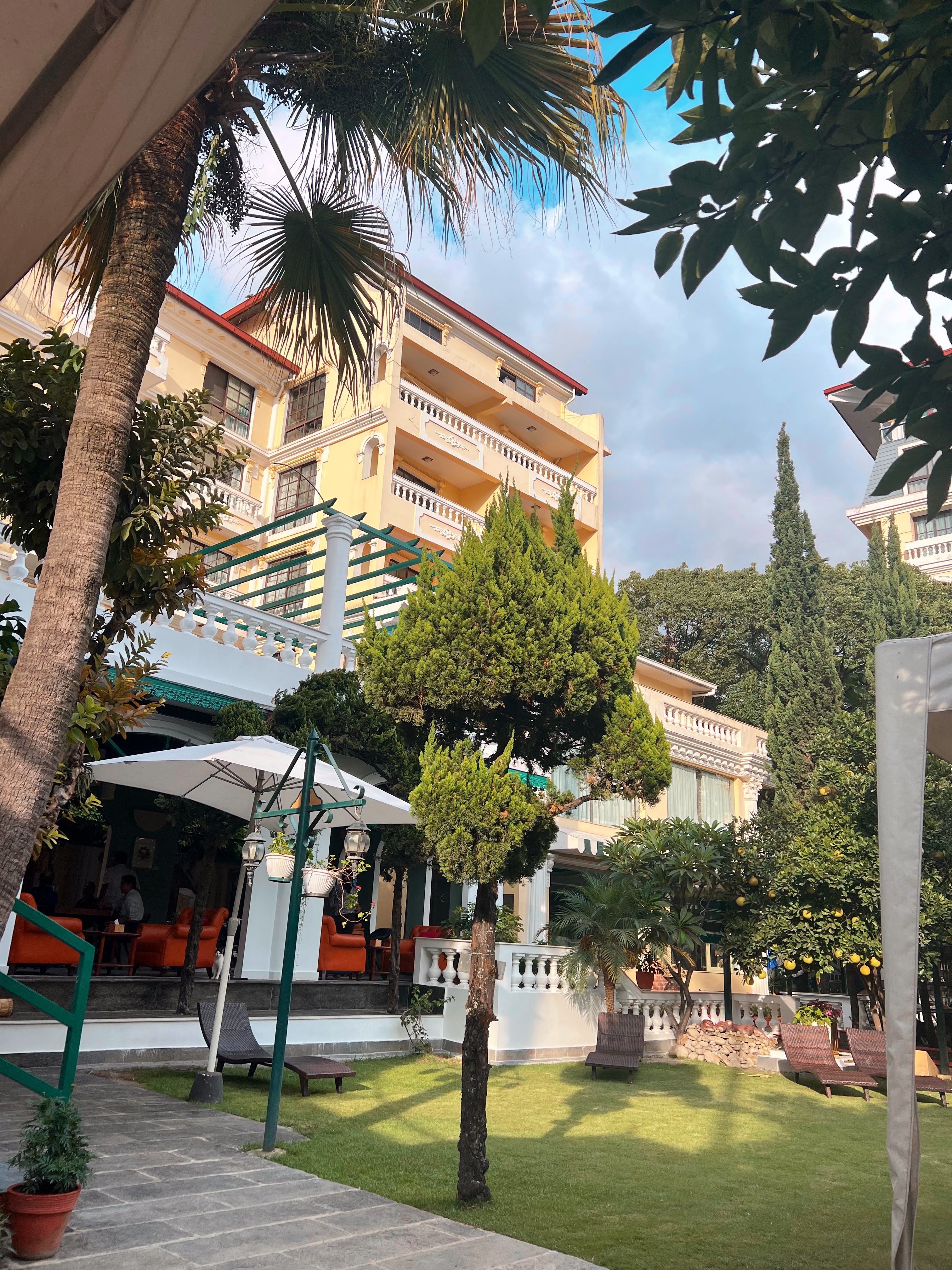 A large, multi-story yellow building with balconies stands behind a green lawn, some trees, and an outdoor patio area with lounge chairs and umbrellas.